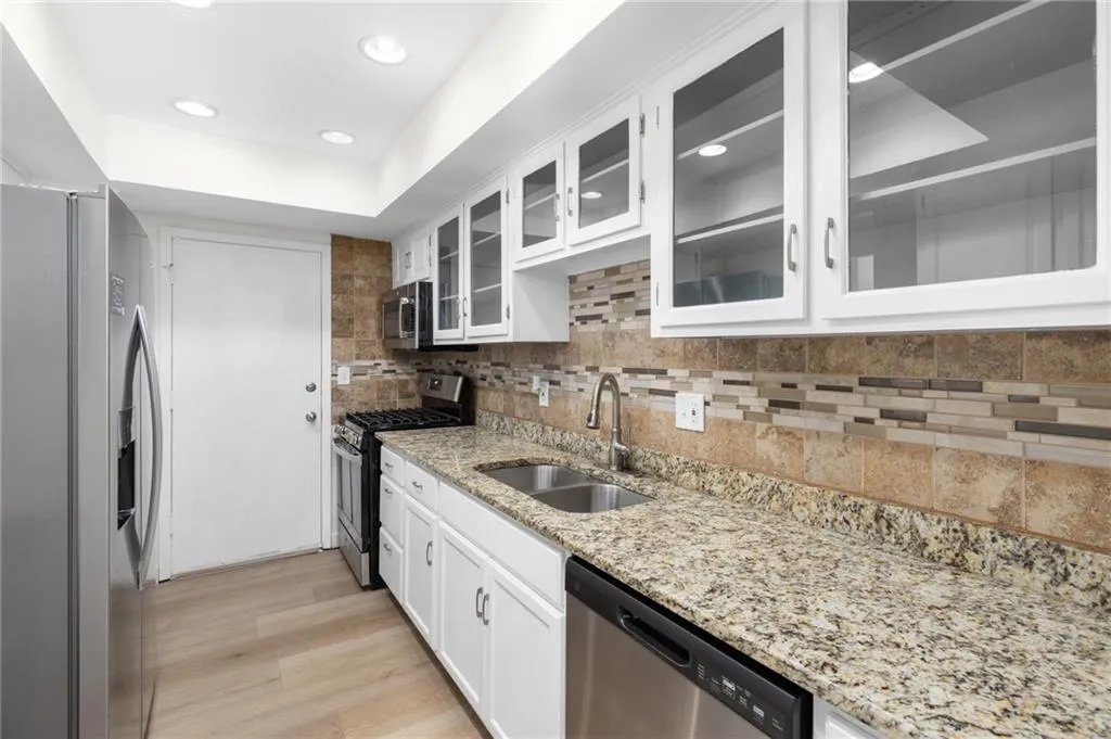 Kitchen featuring glass insert cabinets, stainless steel appliances, white cabinets, light stone counters, and decorative backsplash