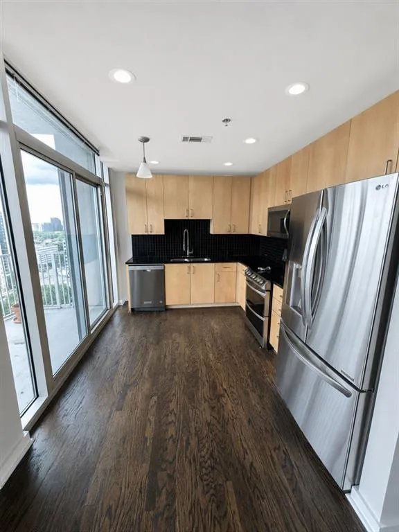 Kitchen featuring dark wood-finish flooring, light wood cabinetry, stainless steel appliances, black tile backsplash, and a sliding glass door to an outdoor space