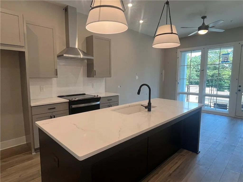 Kitchen featuring wall chimney range hood, black range with electric cooktop, decorative backsplash, light wood-style floors, and a center island with sink