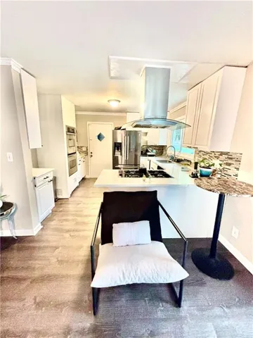 Modern kitchen featuring white cabinetry, a central island with a gas cooktop, a stainless steel range hood, built-in double ovens, and wood-finish flooring