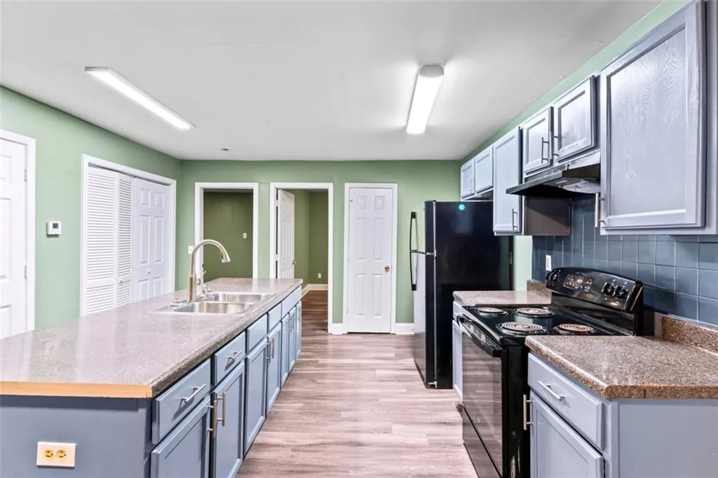 Kitchen featuring black range with electric cooktop, under cabinet range hood, light wood-style floors, decorative backsplash, and a kitchen island with sink