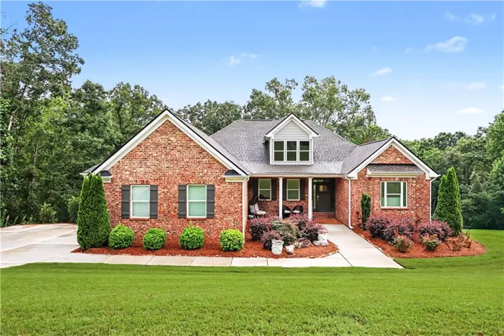 Brick Colonial home featuring a gabled roofline with dormer windows, manicured front landscaping with flowering shrubs and evergreen plantings, circular driveway, and a welcoming front entry with covered portico