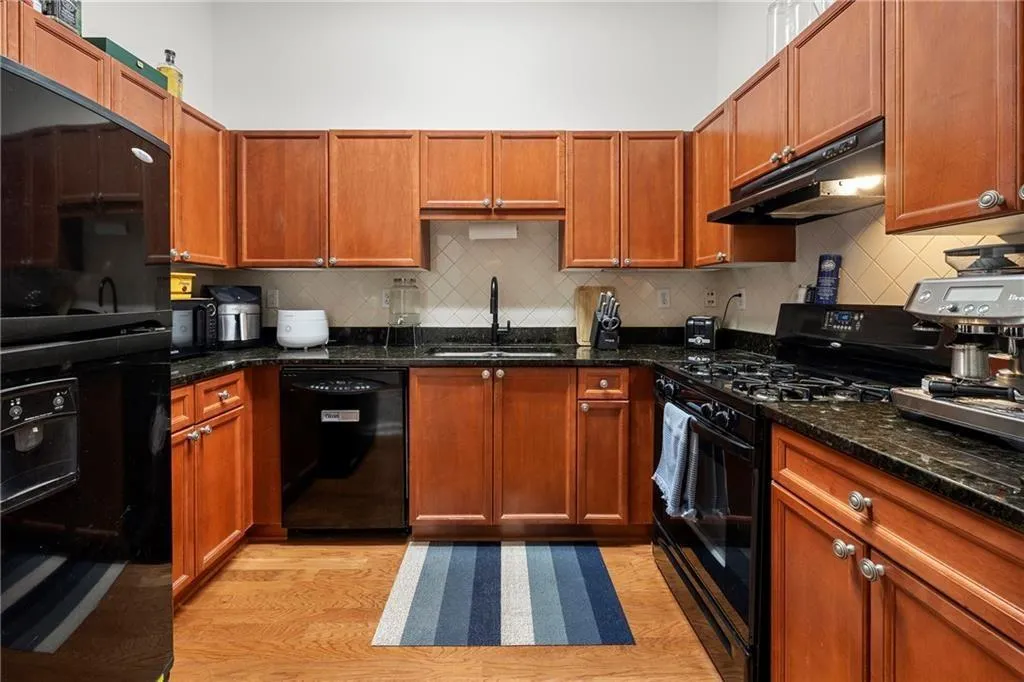 Kitchen with black appliances, light wood-style flooring, under cabinet range hood, tasteful backsplash, and brown cabinets