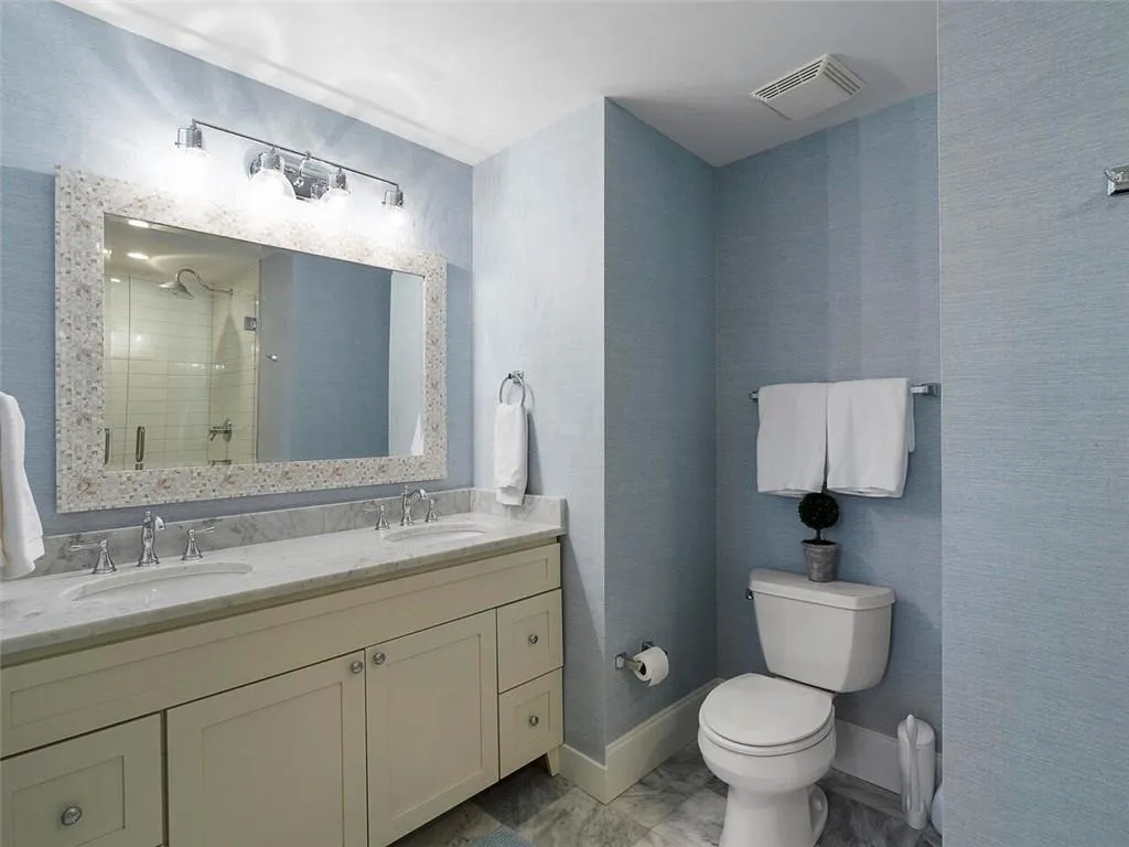 Dual vanity with undermount sinks and chrome faucets, marble-finish countertop, light wood-finish cabinetry with silver-tone hardware, and a framed mirror with integrated lighting