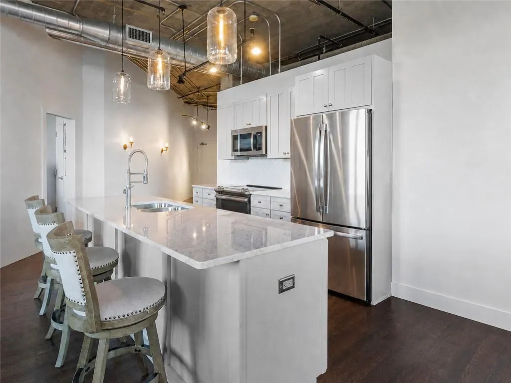 Open-concept kitchen featuring an expansive island with a built-in sink, stainless steel appliances, white shaker cabinetry, and industrial-style exposed ductwork and ceiling