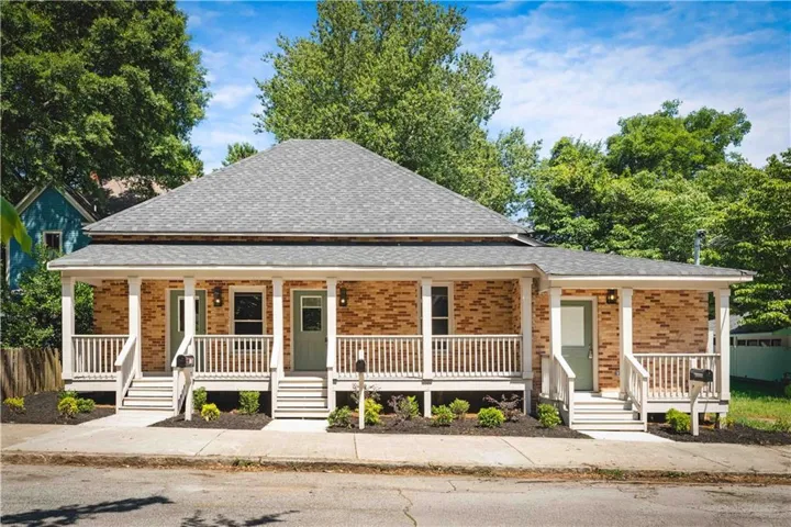 Brick exterior with gray shingle roofing, multiple covered front porches, white railing, and decorative landscaping