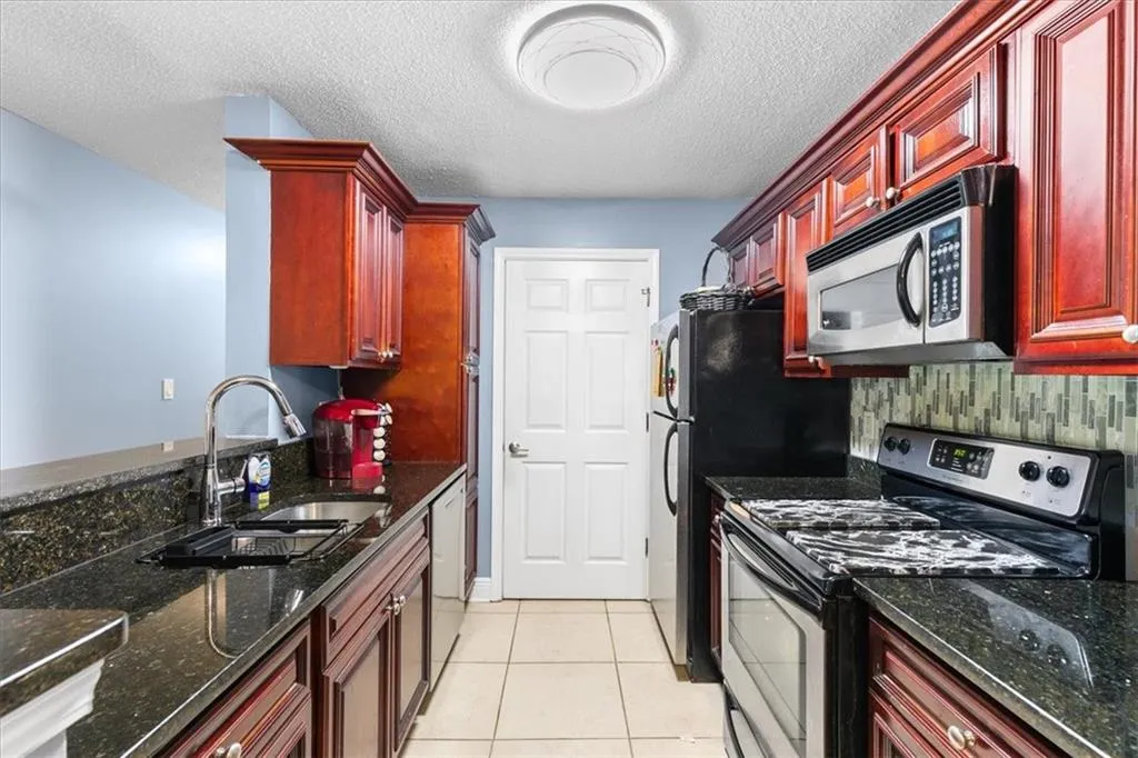 Kitchen with stainless steel appliances, light tile flooring, a textured ceiling, dark stone counters, and sink
