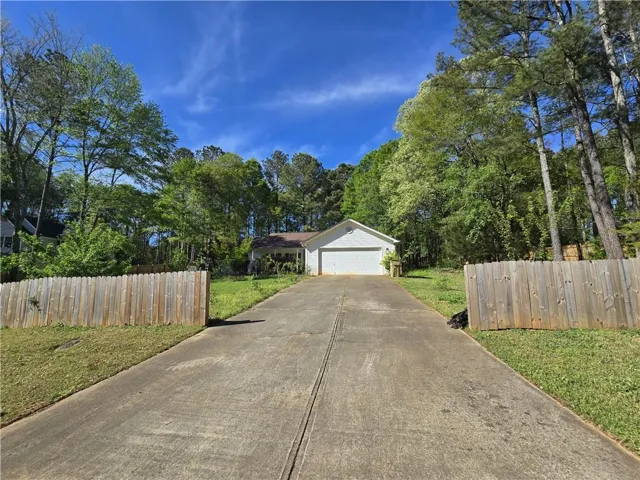 Gated property entrance with a wood fence, concrete driveway, and a single-story residence featuring an attached garage and mature trees