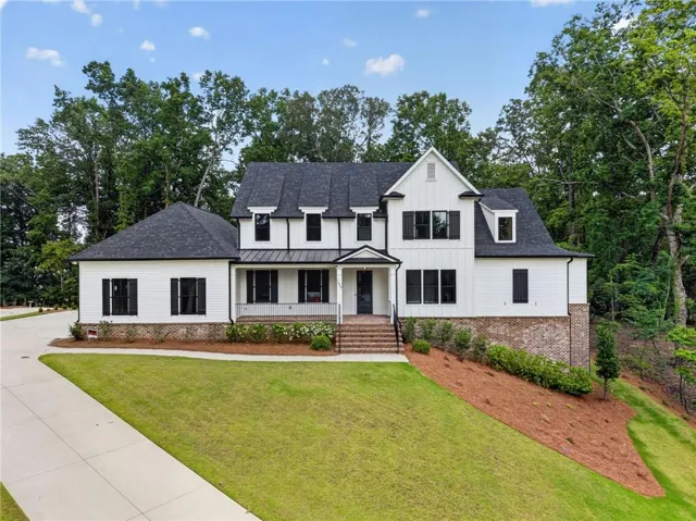 Modern farmhouse architecture featuring white siding, dark shingle roof, black window shutters, brick foundation, and a concrete driveway