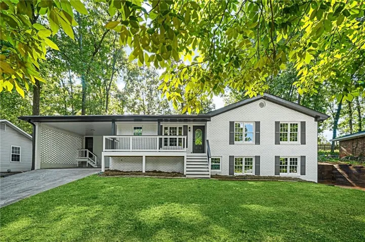 Split-level residence featuring a white brick exterior, dark window shutters, an elevated white deck with black railing, a covered carport with a decorative screen, and a separate shed structure