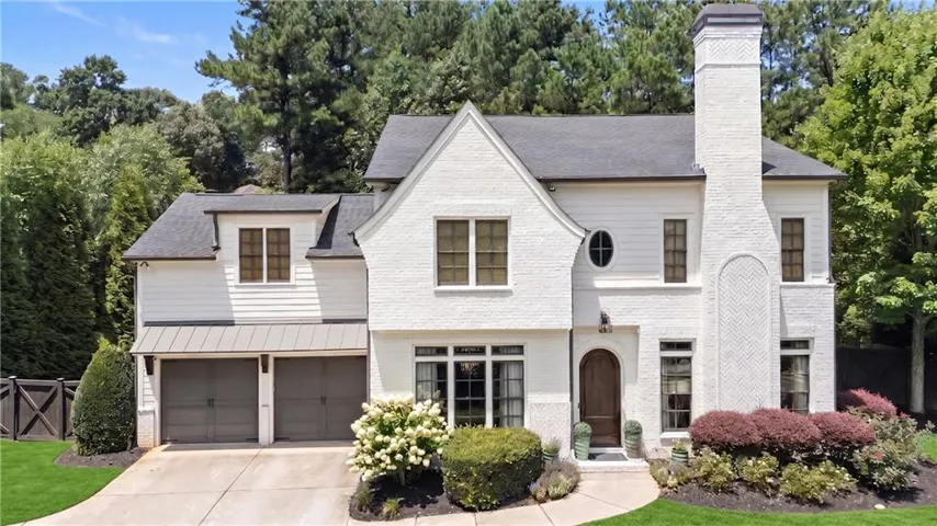 Traditional-style residence featuring a white brick and horizontal siding exterior, a prominent chimney with herringbone detail, a two-car garage with a metal awning, an arched wooden front door, and mature landscaping
