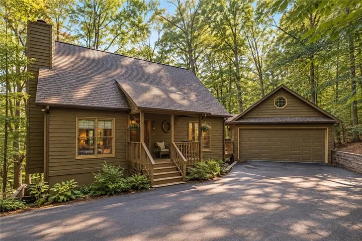 Wooded property with a gabled roofline, covered front porch, attached garage, and paved driveway