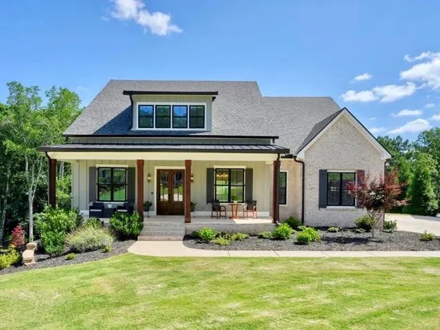 Gable-style roofline with dormer windows, covered front porch with dark wood pillars, light-colored brick and vertical siding exterior, and a manicured lawn