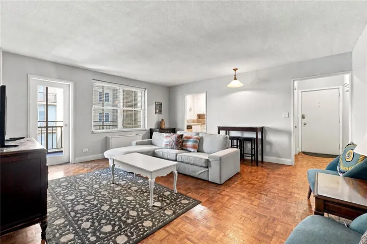 Parquet wood flooring, a window with horizontal blinds, a white door leading to an exterior balcony with black metal railing, a hanging ceiling light fixture, and a white entrance door