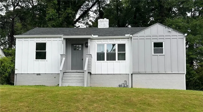 View of front of home featuring board and batten siding, roof with shingles, a front lawn, a chimney, and view of wooded area