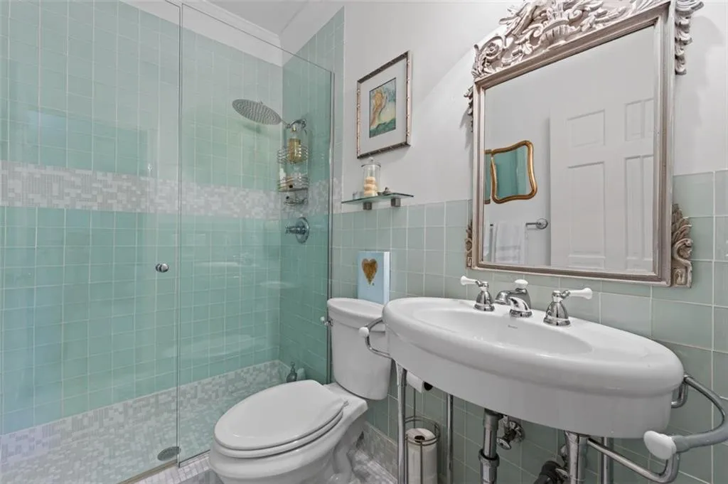 Glass-enclosed shower with a rain showerhead, light blue tiled walls featuring a mosaic accent band, decorative framed mirror, and a pedestal sink with chrome fixtures