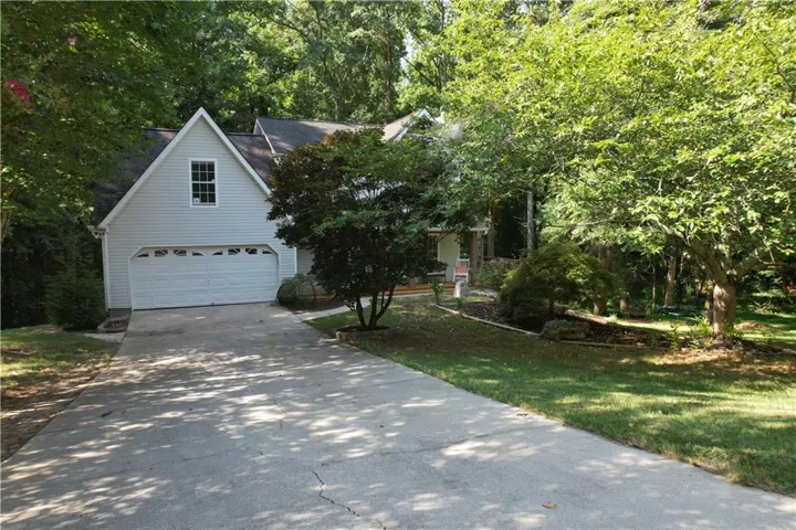 Exterior featuring light-colored siding, a two-car garage, an elongated concrete driveway, and mature landscaping with deciduous trees