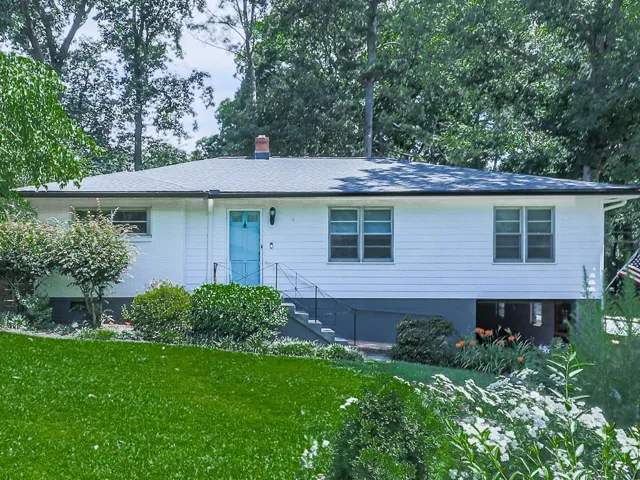 White siding exterior with a light blue entry door, featuring a black shingle roof, dark gray foundation, and established landscaping