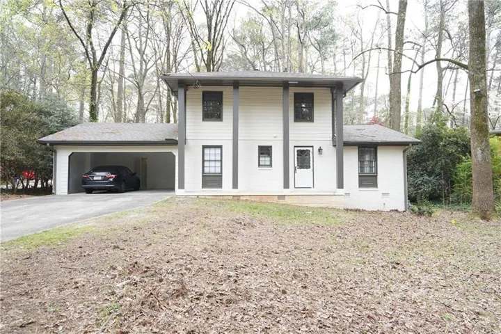Two-story residence featuring a covered carport, white exterior siding, dark trim accents, a prominent front entry with a white door, and surrounding mature trees