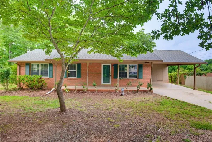 Brick ranch home featuring a welcoming front entrance with dark green door and shutters, expansive driveway with carport, and mature shade trees throughout the landscaped grounds