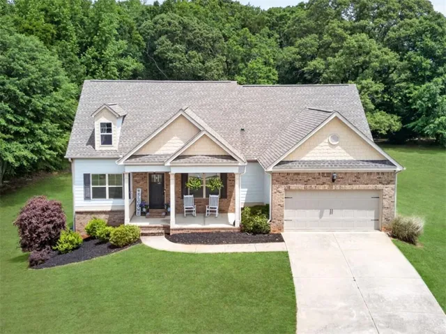 Charming brick and siding cottage with covered front porch, featuring a two-car attached garage, gabled roofline with architectural detail, manicured landscaping with mature shrub plantings, and expansive level lawn bordered by established trees