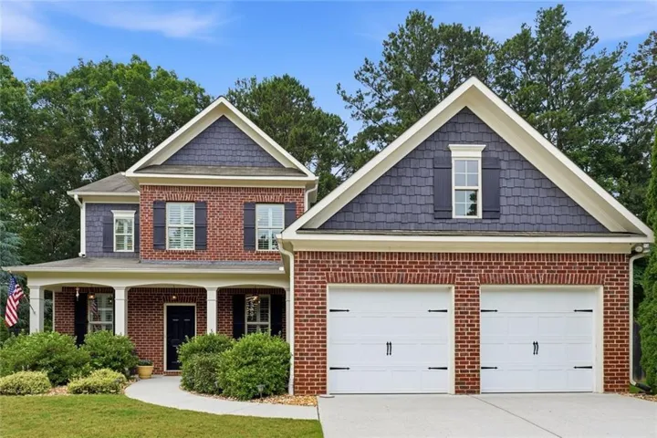 Traditional brick facade residence featuring a covered front porch with white columns, dark shutters, a two-car garage, and a curved concrete walkway