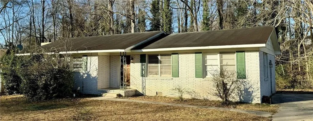 White brick exterior with green window shutters, a dark shingle roof, and a mature tree line backdrop
