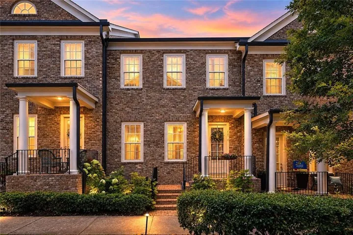 Brick exterior with white-framed windows, columned entryways, manicured hedges, and exterior lighting