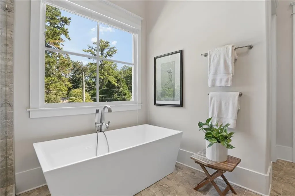 Freestanding soaking tub with a chrome handheld shower fixture, large window, and tile flooring