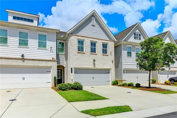 Traditional townhome featuring clean-lined architectural design with brick and siding exterior, multiple gabled rooflines, two-car attached garage with white paneled doors, expansive concrete driveway, and manicured front landscaping with established lawn and planted beds
