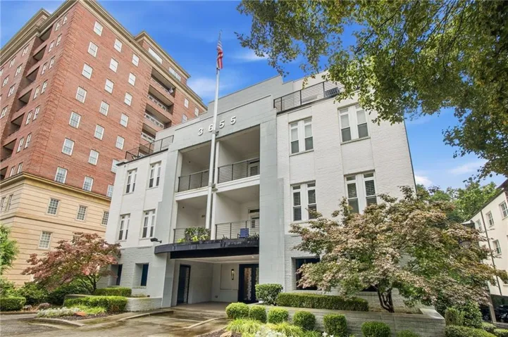 Contemporary multi-story building featuring a prominent flagpole, multiple balconies with dark railings, a covered drive-through entrance, and mature landscaping with blooming trees