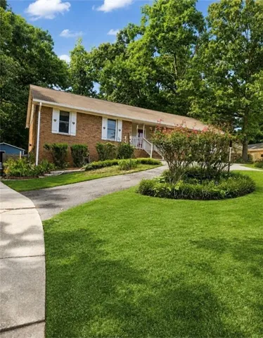 Brick exterior, front porch with white railing, paved driveway, mature trees, and landscaped shrubbery