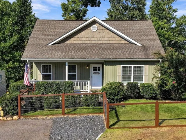 Gable roofline with architectural shingles, front porch with white railings, natural wood-tone privacy fence, gravel driveway, and exterior siding with shake accents