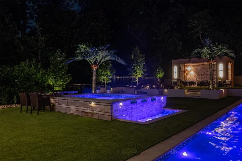 Luxurious outdoor entertaining space featuring an illuminated resort-style pool with integrated spa, artificial turf, palm trees, ambient landscape lighting throughout, and adjoining poolside cabana
