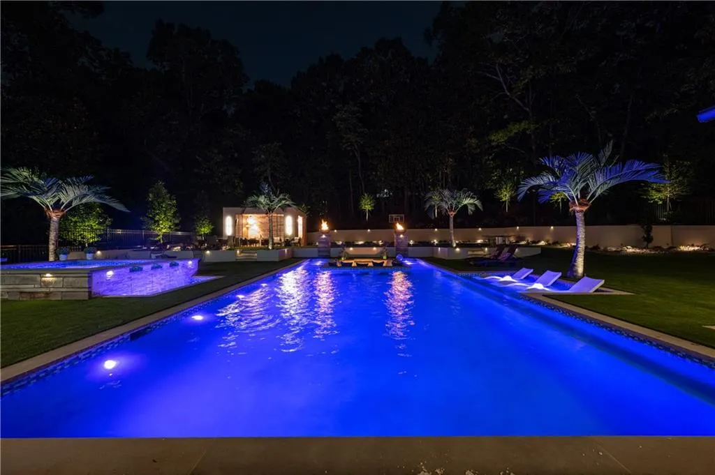 Luxurious outdoor entertaining space featuring an illuminated resort-style pool with integrated spa, artificial turf, palm trees, ambient landscape lighting throughout, and adjoining poolside cabana