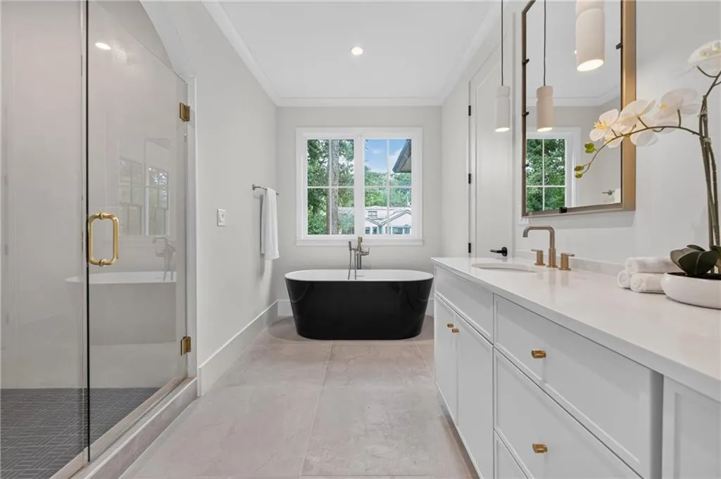 Spacious bathroom featuring a frameless glass shower enclosure, a freestanding black soaking tub, a double vanity with white cabinetry, a large window, and light-toned tile flooring
