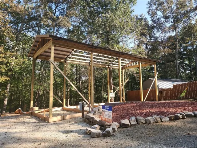 Open-air wooden structure with a pitched roof, gravel surfacing, and a stone-edged landscape bed