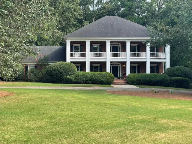 Traditional brick colonial residence featuring a two-story white columned portico with railing, a dark shingle roof, and a mature landscaped front yard with established shrubbery