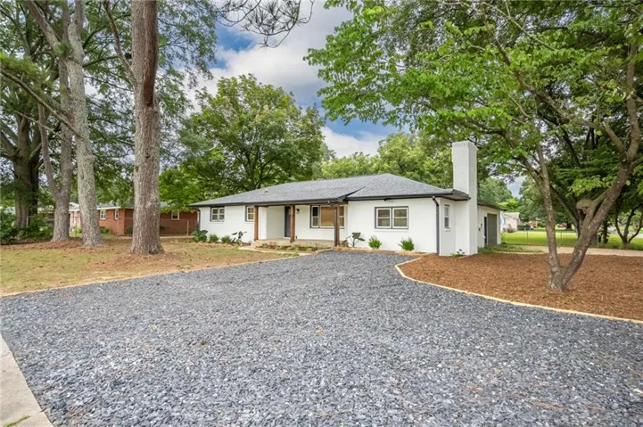 Exterior featuring white siding, a dark roof, a gravel driveway, and mature landscaping