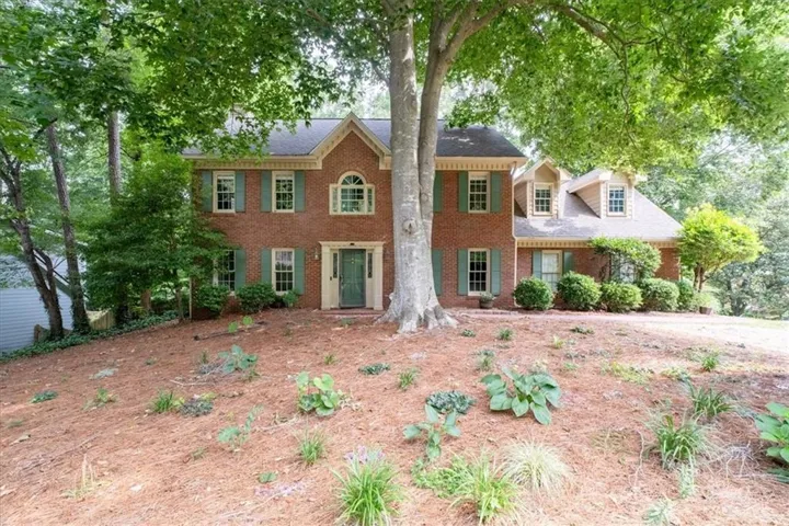 Traditional brick residence featuring a prominent mature tree, symmetrical window placement, decorative green shutters, a central entrance with a green door, and dormer windows on the upper level