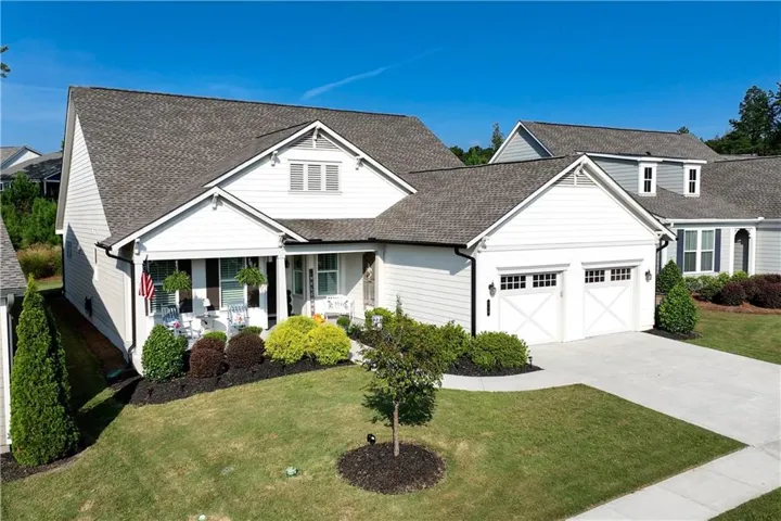 White siding exterior with a dark shingle roof, covered front porch, attached two-car garage with carriage-style doors, and concrete driveway