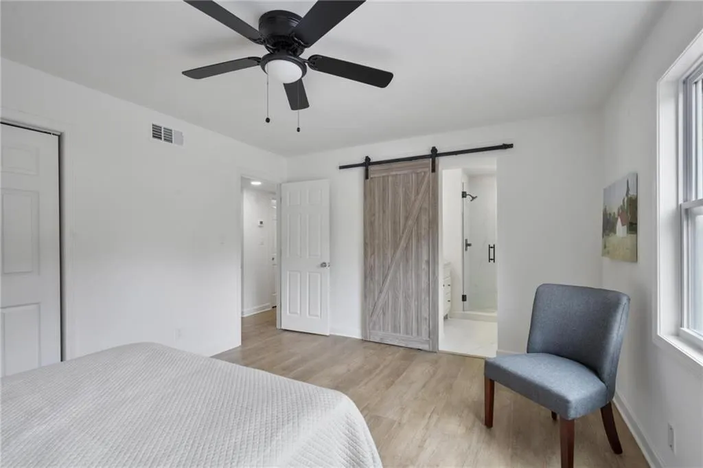 Bedroom with light wood-type flooring, ensuite bathroom, a barn door, and ceiling fan