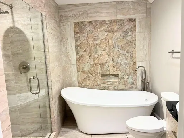 Freestanding soaking tub, floor-mounted tub filler, stone-look accent wall, tiled shower with glass enclosure, and a rain shower head