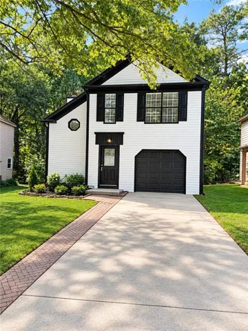 White siding exterior with black trim, an attached garage, a concrete driveway with a brick paver border, and mature landscaping