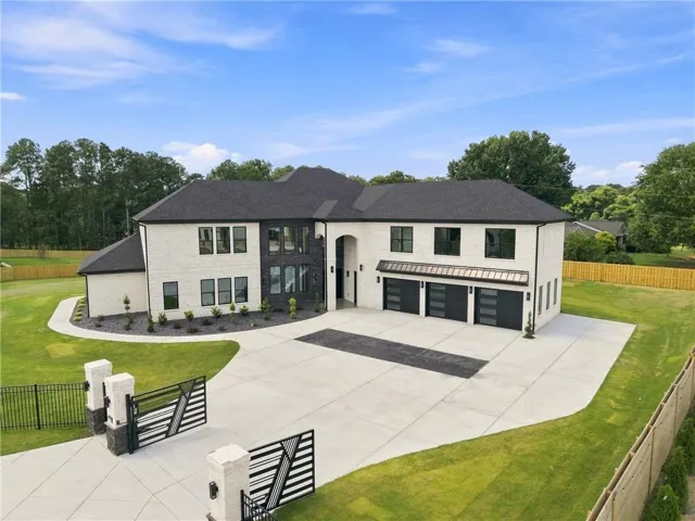 White brick facade with dark window frames, three-car garage with modern doors, gated entry, extensive concrete driveway, and a dark shingle roof