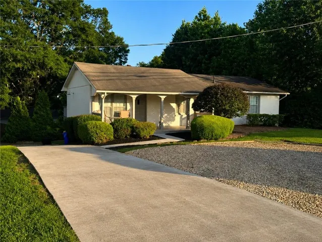 Charming single-story home featuring a covered front porch with decorative columns, concrete driveway, and mature landscaping with established shrubs and trees