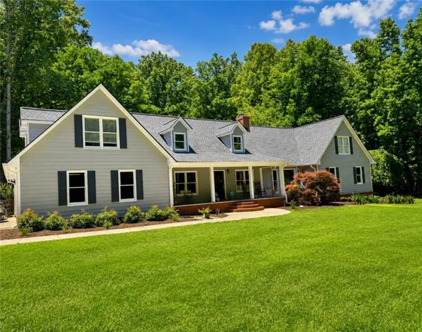 Expansive front lawn with a light gray siding residence featuring a covered porch, multiple dormer windows, dark shutters, and a brick chimney