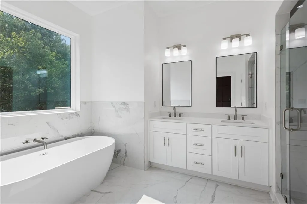 Freestanding soaking tub with wall-mounted faucet, dual vanities with quartz countertops, framed mirrors, and glass-enclosed shower