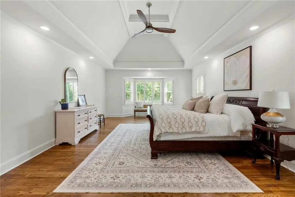 Spacious primary bedroom with vaulted trey ceiling, hardwood floors, and abundant natural light. A cozy sitting area by the bay windows with plantation shutters creates a serene retreat.