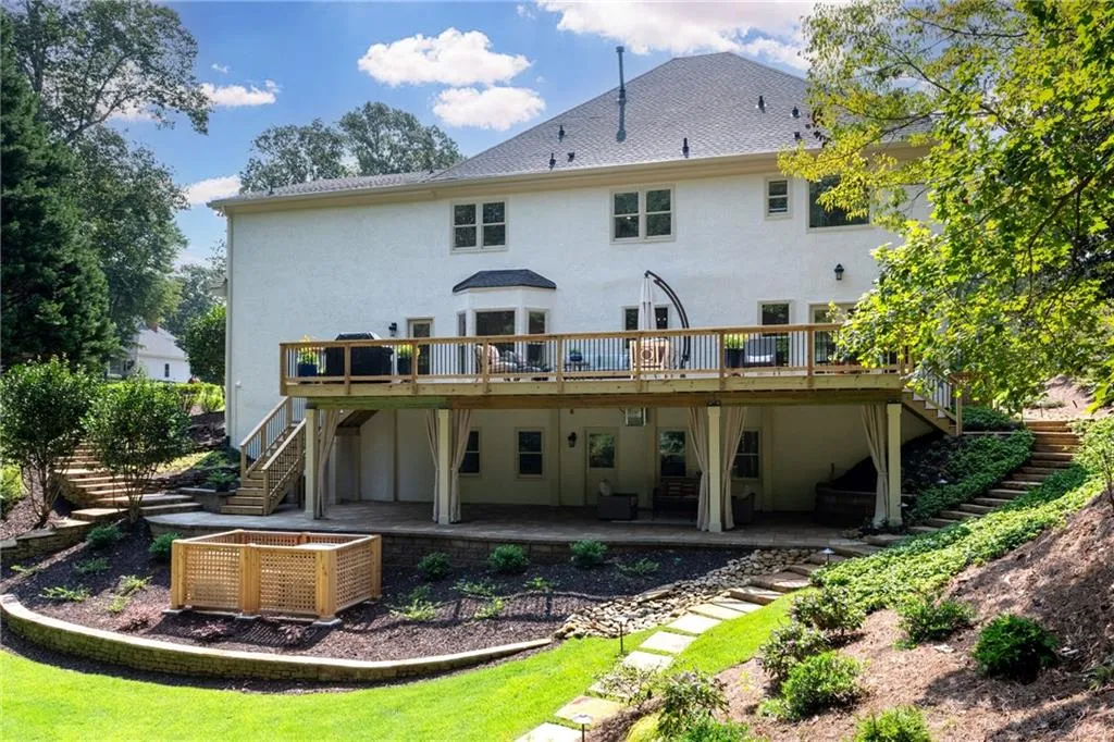 Beautiful rear exterior featuring an expansive elevated deck, covered paver patio below, landscaped terraced yard, and manicured lawn with raised garden bed perfect for entertaining and outdoor living. At night, the backyard comes alive with path and deck lighting.
